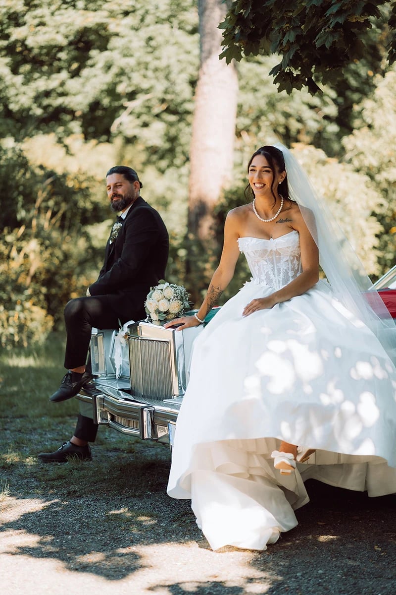 Braut sitzt auf der Motorhaube des Hochzeitsautos, Bräutigam daneben — Hochzeit Elisabete & Nafi in Hof